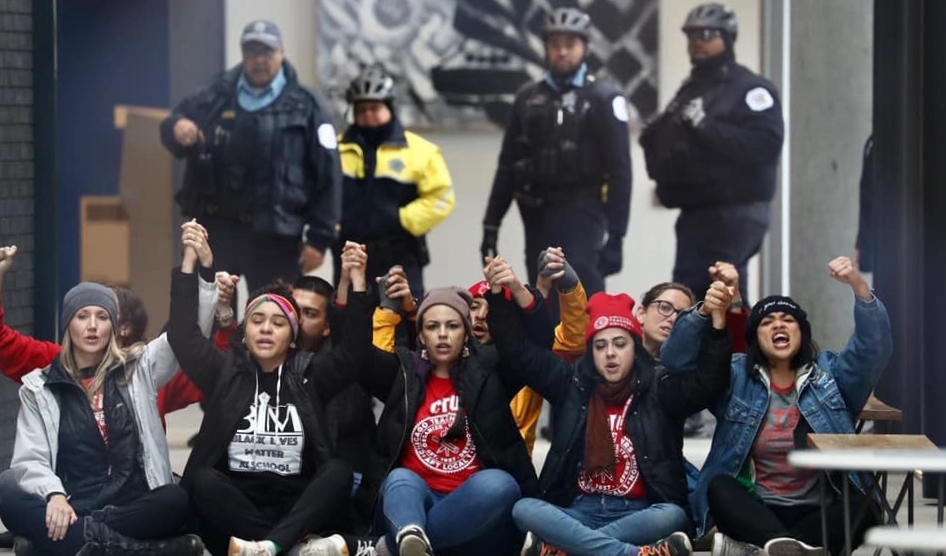 Teachers seated on sidewalk with joined hands raised; police standing behind them