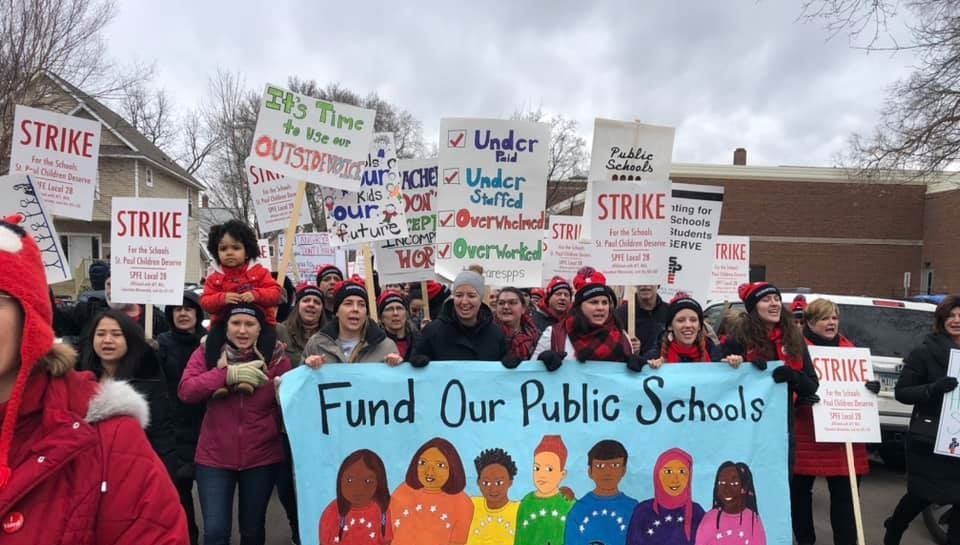 Strikers carry a banner "Fund Our Public Schools" with a rainbow of faces. Picket signs say "Strike" and "It's Time to Use Our Outside Voice." One kid is carried on an adult's shoulders.
