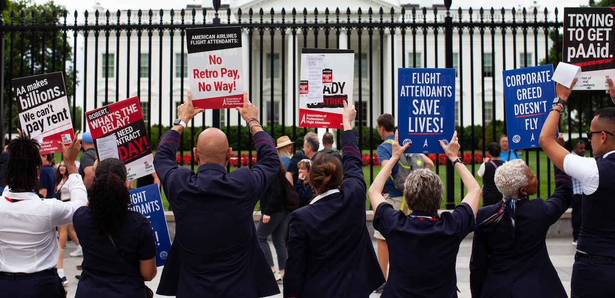 A line of seven flight attendants in uniform, viewed from behind, stands at the White House gates holding up various printed signs. Messages say: "AA makes billions, we can't pay rent." "Robert, what's the delay? We need a contract today!" "American Airlines flight attendants: No retro pay, no way!" "Flight attendants save lives!" "Corporate greed doesn't fly." "Trying to get pAAid." It appears the group includes men and women, and most are people of color.