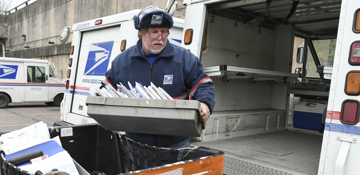 A man with a white beard in a blue postal uniform loads a tray of letters in to the back of a postal truck.