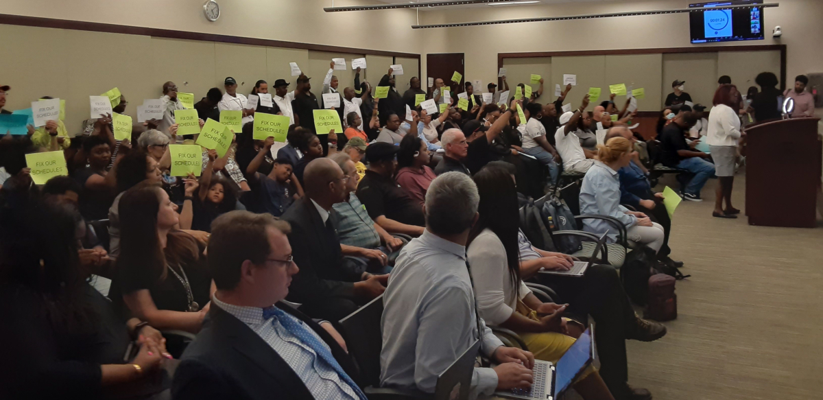 Inside a board meeting, dozens of union members and supporters hold up small yellow and white signs reading 'Fix Our Schedules.'