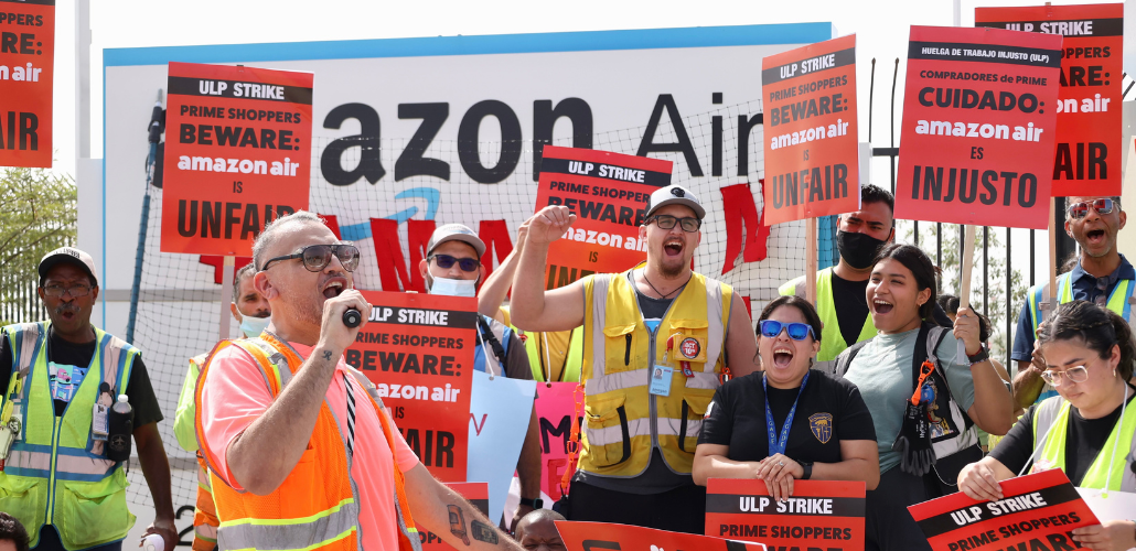 Workers in orange vests yell together, outdoors in front of an Amazon Air sign. Person in front has a microphone. Several have red printed picket signs saying "ULP strike. Prime shoppers beware: Amazon Air is unfair." Or the same in Spanish.