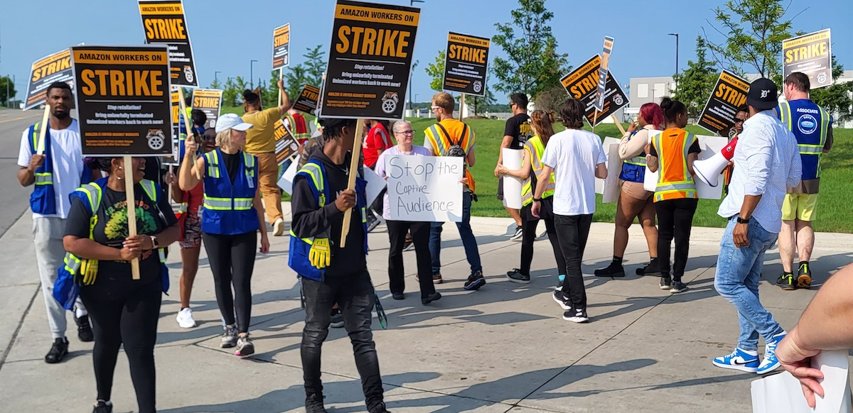Workers (men and women, Black and white) picket outside in the sunshine. Some carry printed signs that say "Amazon workers on strike" with a Teamsters logo. One carries a hand-written sign that says "Stop the captive audience."