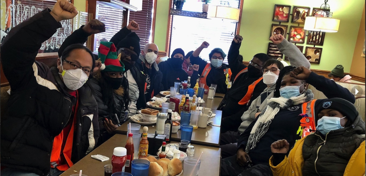 A group of workers sitting at a long restaurant table, some in safety orange vests and wearing surgical masks, raise their fists and look at the camera.