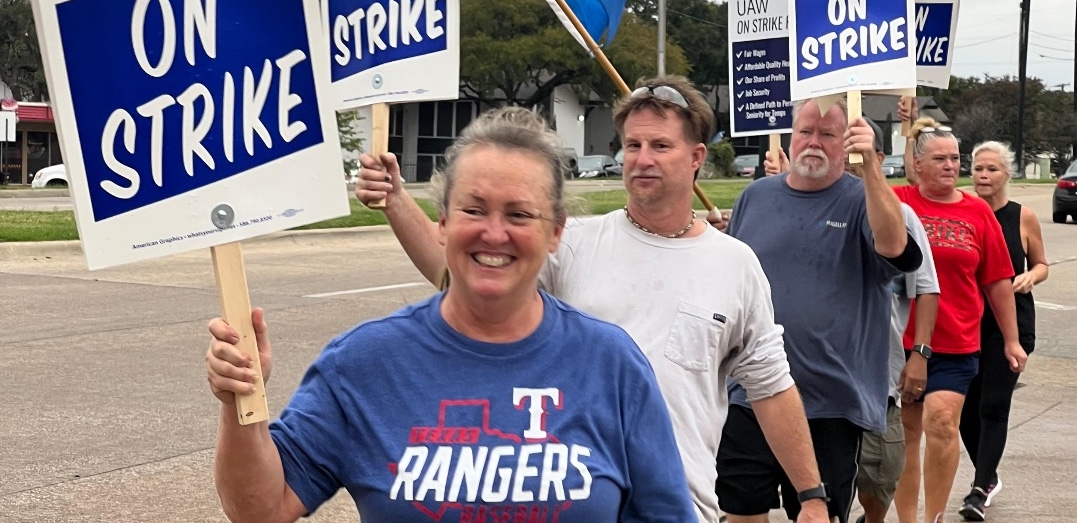 A woman with a blue Texas Rangers T-shirt and a sign that says “On Strike” smiles as she leads a line of autoworkers towards the camera.