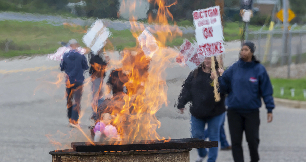 Strikers march holding picket signs that read "On Strike," while a fire rises from a burn barrel to keep them warm.