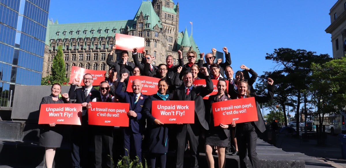 20 flight attendants in uniform hold red signs saying “unpaid work won’t fly” in both French and English