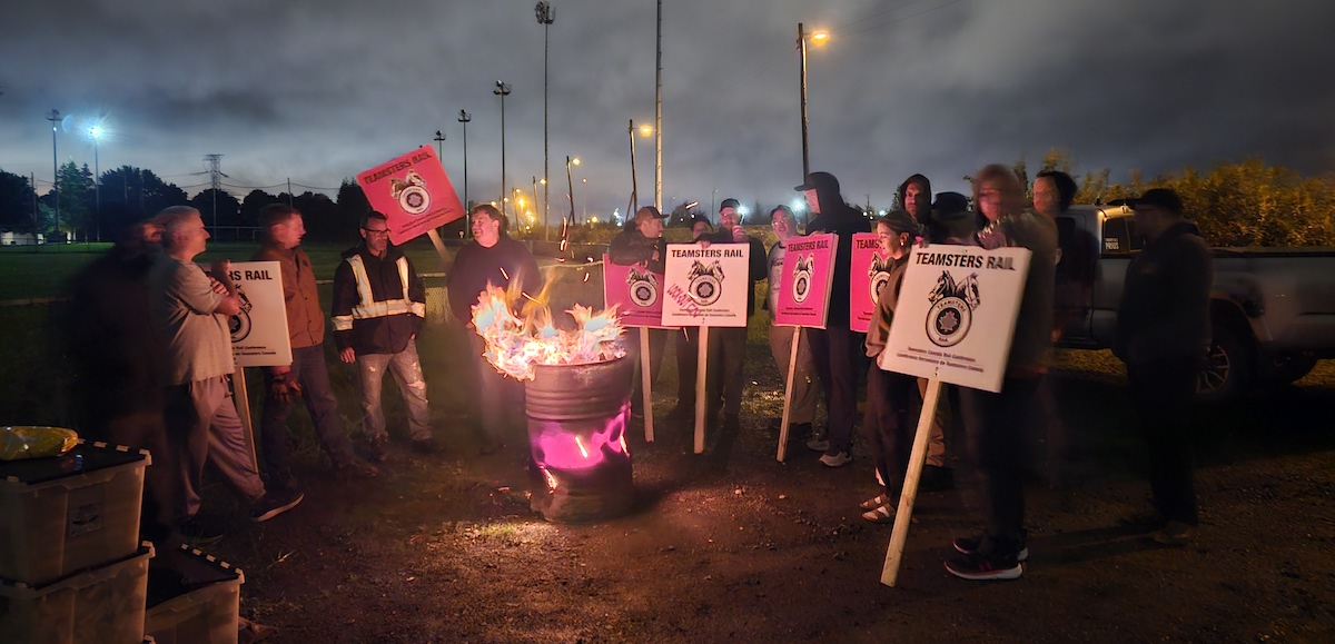 Around dawn, but still dark, a group stand around a burn barrel with signs saying Teamsters Rail locked out