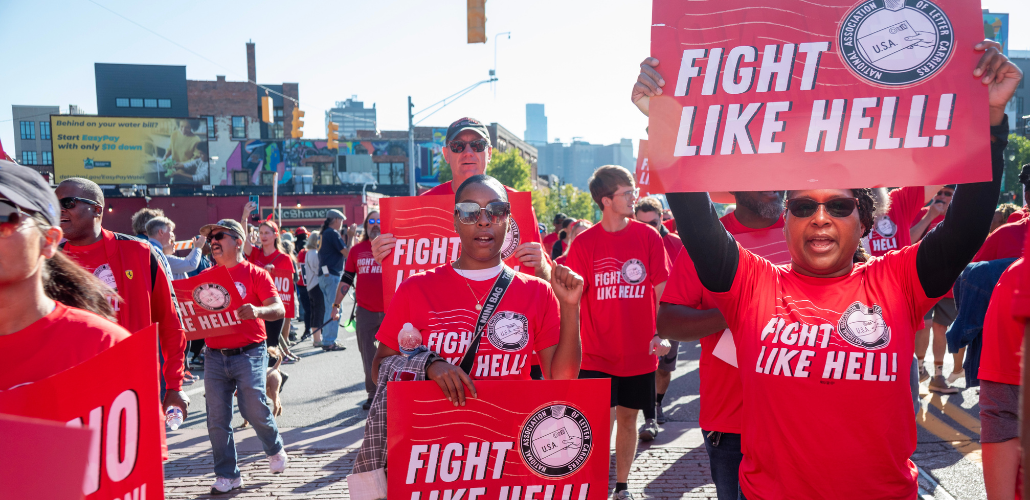 Black and white women and men march through a Detroit street. Everyone is wearing a red "fight like hell" T-shirt with the NALC logo, and some people are carrying printed signs with the same design. Most prominent in the frame, looking directly into the camera, are two Black women in sunglasses, one raising a fist at shoulder level and the other holding a sign above her head.