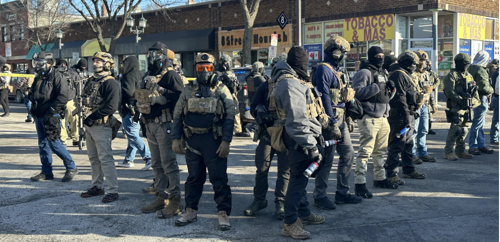 Agents covered head to toe in tactical gear and warm clothing stand in a diamond formation, facing outward, in a city street. Their faces are covered. The vests say "police" though they are not in uniform. They all wear heavy boots and some carry spray cans. Visible behind them is a one-story brick building lined with retail shops, with faded signs reading "8 Street Nourish" and "Tobacco & Mas."