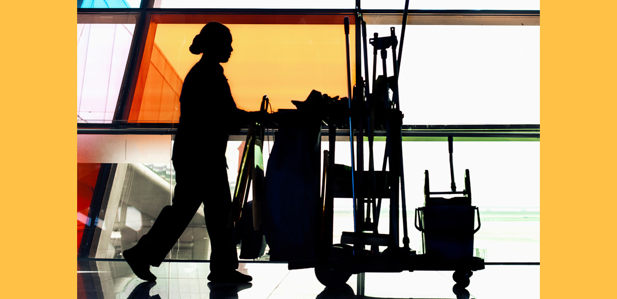 A woman custodian pushes a cart full of implements in profile past colorful windows.