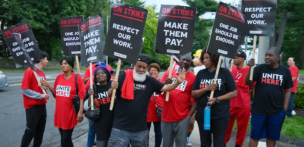 A group of a dozen hotel workers with signs saying “On Strike, Make them Pay” look at the camera, one waves