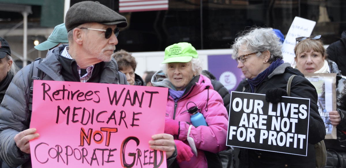 Three older people in a crowd hold signs saying “Our lives are not for Profit” and “Retirees want Medicare, Not Corporate Greed”