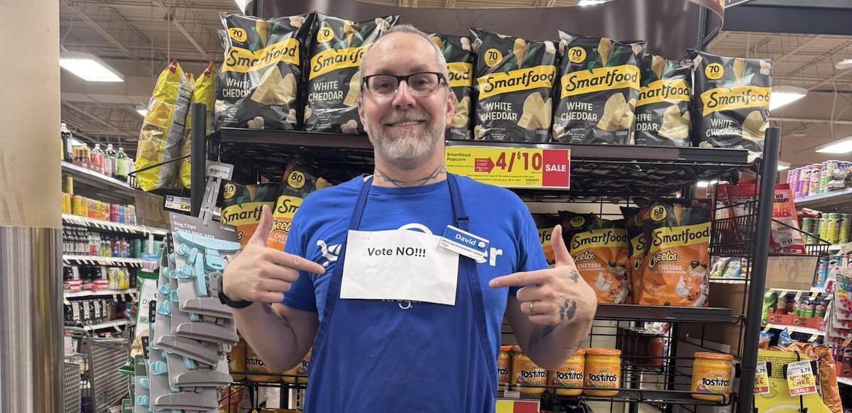 A man in in front of grocery shelves points to a printed-out “Vote No” sign pinned to his blue shop apron.