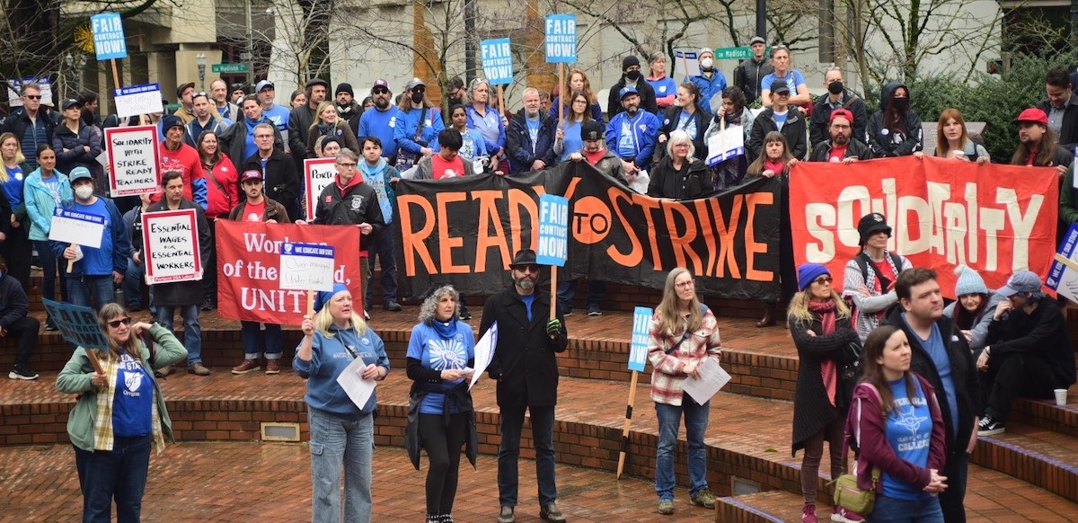 People rally on brick steps (Portland's Pioneer Courthouse Squre) on a rainy day. Many wear blue or red T-shirts under their coats. Banners say "Ready to strike" and "Soldarity." Picket signs say "Fair Contract Now," "Solidarity with Strike-Ready Teachers," and "Essential Wages for Essential Workers."