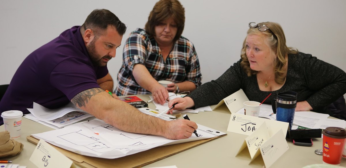 Three people reach across a table to mark a drawing of their workplace