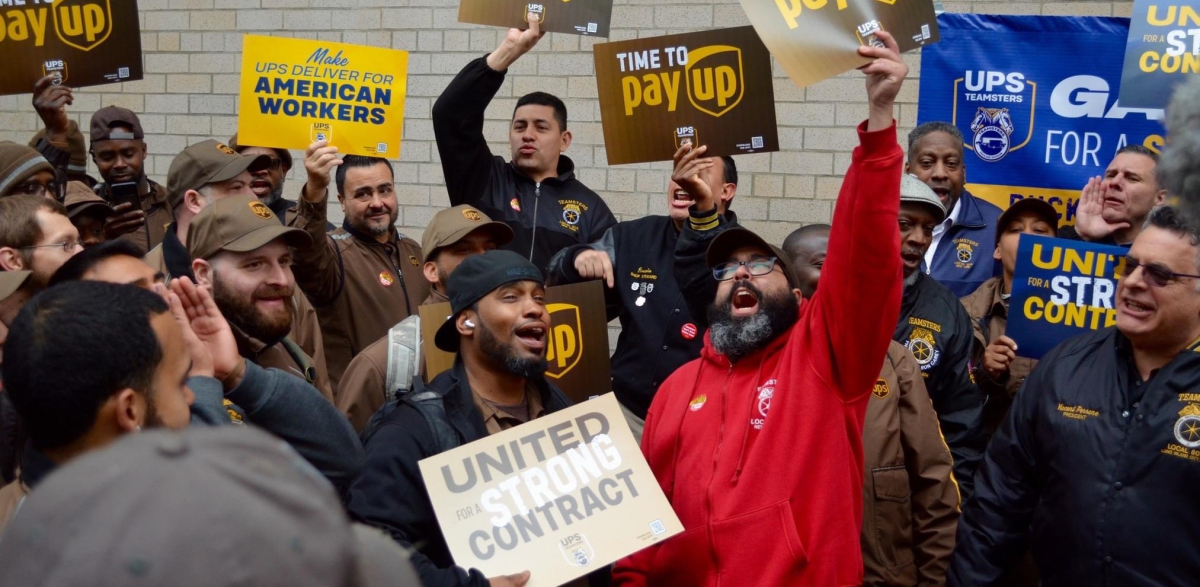 A group of excited Teamsters, some in brown uniforms, others in union hoodies,  hold signs saying time to pay up,