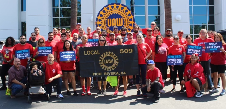 A group of 30 red-shirted autoworkers stand around a banner and hold contract campaign signs.