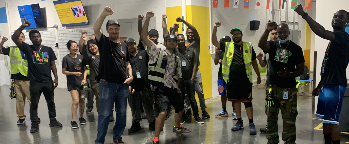 A group of smiling young workers, diverse in race and gender, raise their fists on the warehouse floor. Their pose is casual and triumphant -- looks like they just came from collective action. Some wear safety vests.