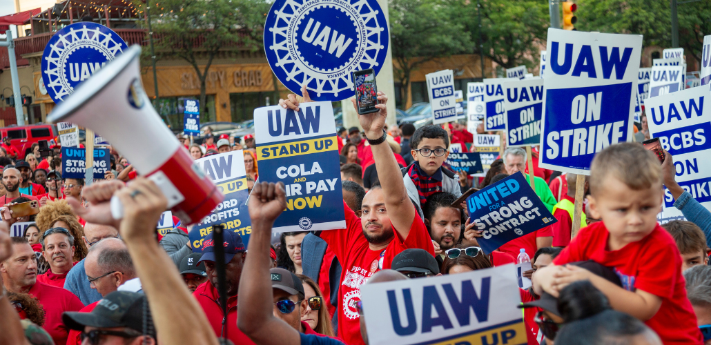 UAW members and supporters at a rally. Many hold signs saying UAW on Strike. Several have their children on their shoulders.