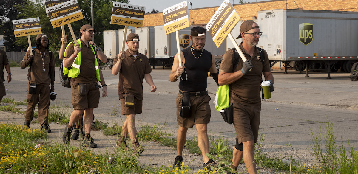 A line of men in brown UPS uniforms and backwards hats with signs saying “Just Practicing for a Just Contract” walk by trailer trucks labeled UPS.