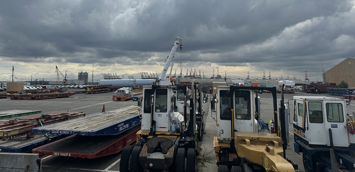 Small port vehicles are in the foreground, and harbor cranes are visible in the distance.