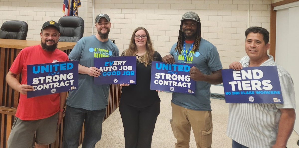 A group of five auto workers pose in their union hall with blue signs that say "End Tiers," "Every Auto Job a Good Job," and "United for a Strong Contract."