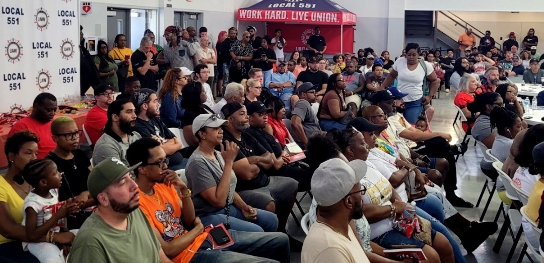 Photo shows a packed hall of autoworkers sitting on folding chairs with standees in the back. 