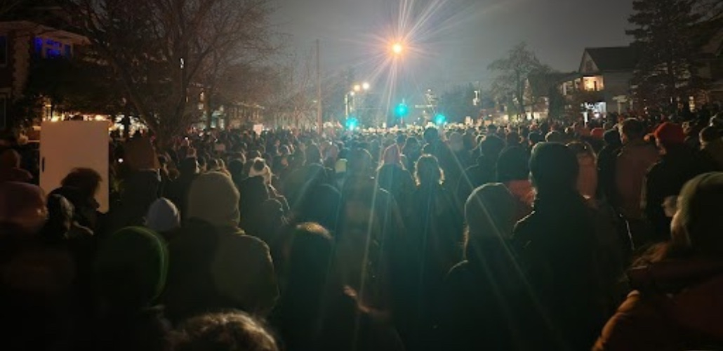 photo taken at night shows a giant crowd of people in a residential street, facing away from the viewer, silhouetted by a streetlight
