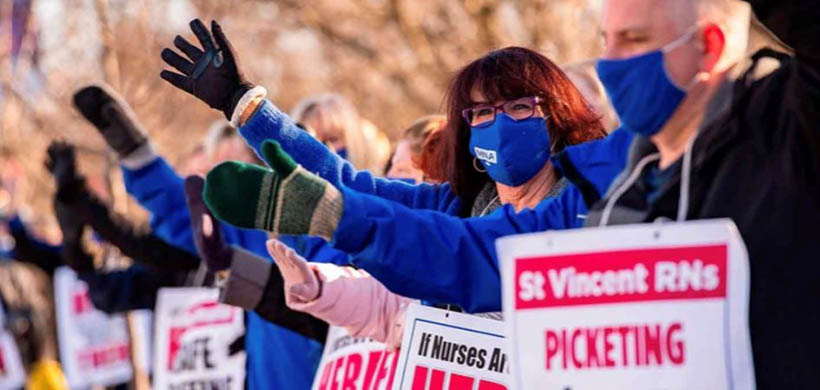 Woman in blue coat facing sideways with hand up among many picketers with hands up, who are holding signs in support of St. Vincent striking nurses