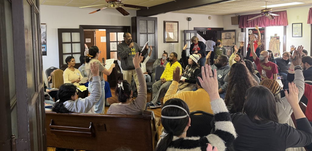A group of people in chairs crowd a room listening to a speaker.