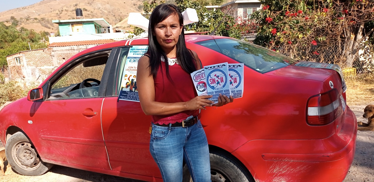 A woman wearing a red blouse and jeans and a serious, determined expression holds up a handful of "VOTA SINTTIA" leaflets, fanned out. She is standing in a sunny outdoor location, in front of a red car that has a poster taped on the side.