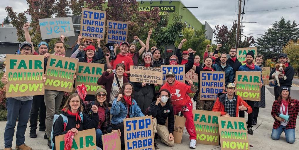 A group of workers and supporters hold cardboard signs reading Stop Unfair Labor Practices and Fair Contract Now. Many are wearing red bandanas.