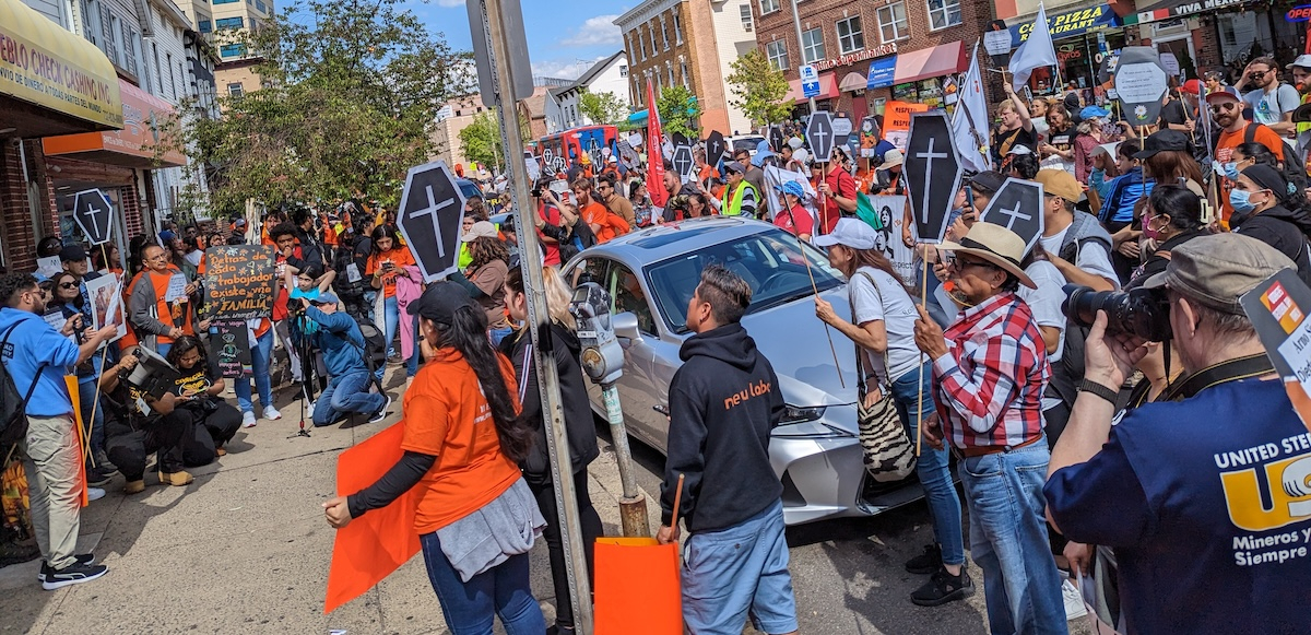 A crowd fills the street holding signs, some of them shaped like coffins.