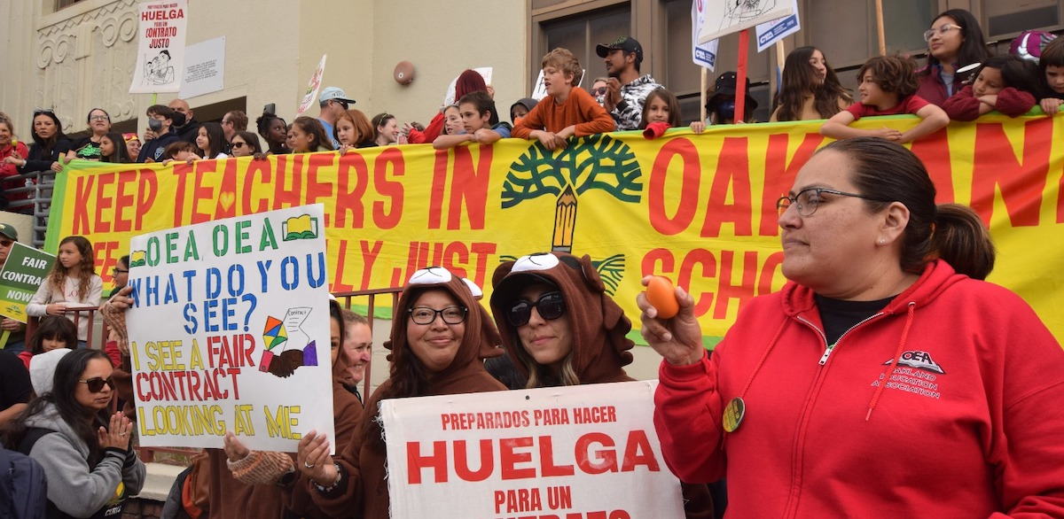 A crowd of adults and children hold a banner saying Keep Teachers in Oakland Schools while other signs say Huelga para un Contracto Justo and Fair Contract Now.