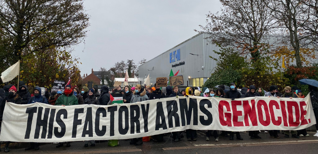  A large crowd of people bundled against the cold, some masked, hold a long banner: "THIS FACTORY ARMS GENOCIDE." A few small handmade signs are also visible: "Ceasefire now, end the occupation," "Jews against genocide," and a Palestinian flag. Behind them is the factory with a big "Eaton" logo.  
