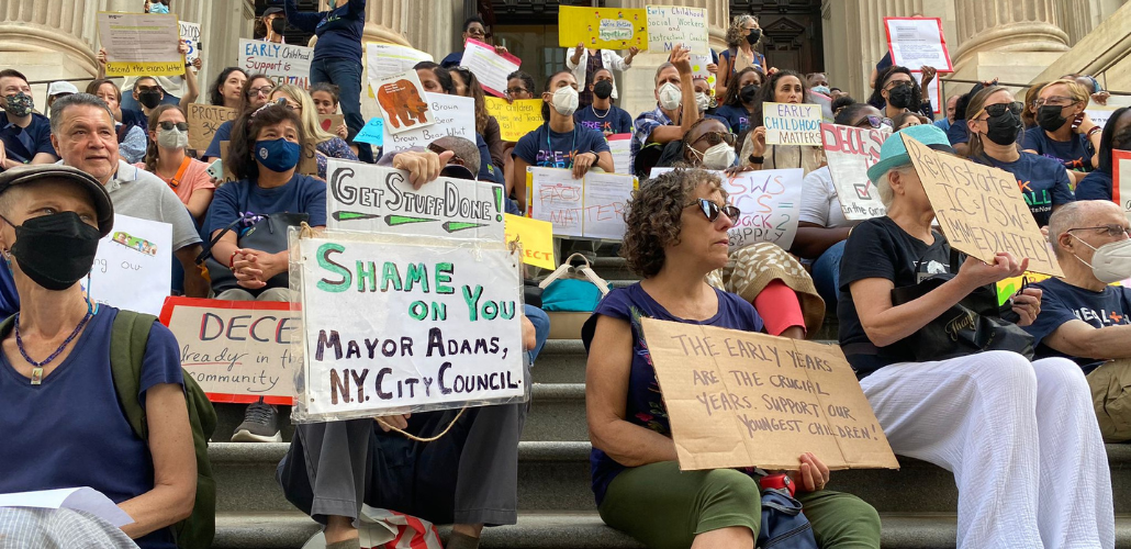 A large crowd of people with handmade signs sit on the steps of a public building. The most visible signs in the foreground say: "Get stuff done! Shame on you Mayor Adams, NY City Council." "The early years are the crucial years. Support our youngest children!" "Reinstate ICs/SWs immediately."