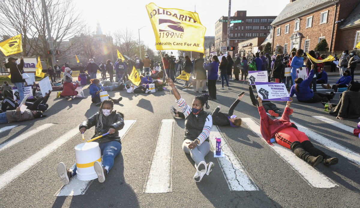 Several women sit in the middle of the crosswalk, holding upside-down buckets that they are using as drums. One holds a flag that reads "SEIU 1199 New England: Health Care for All."