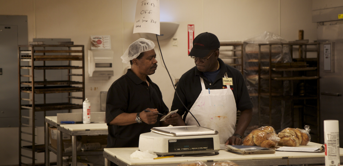 Two bakery workers talk in a kitchen.