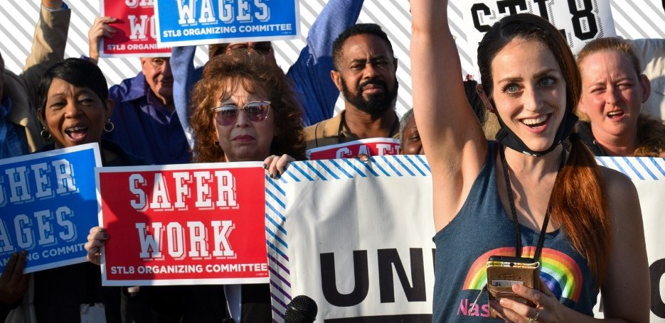 A group of mostly women stand close together with signs saying “Higher Wages” and “Safer Work”