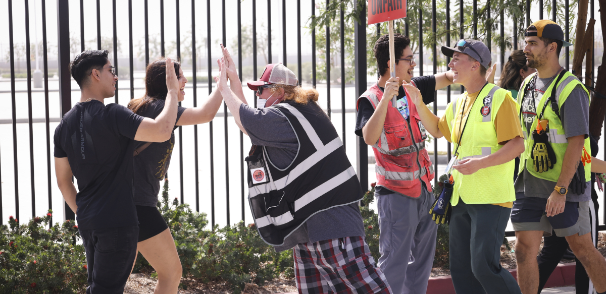 A woman in a safety vest high-fives two others in front of a fence while others stand by with strike signs