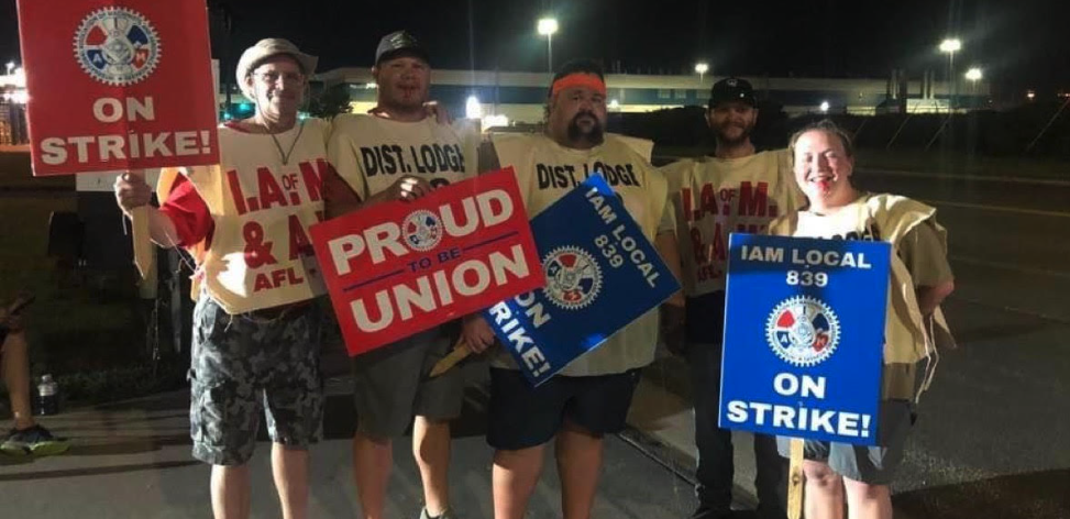 Four men and one woman wearing Machinists tabards and holding strike signs face the camera at night with plant lights behind them.