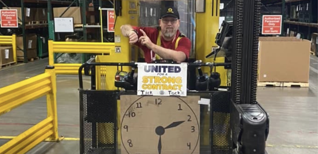 A worker on a forklift points to his watch with a clock sign below him.