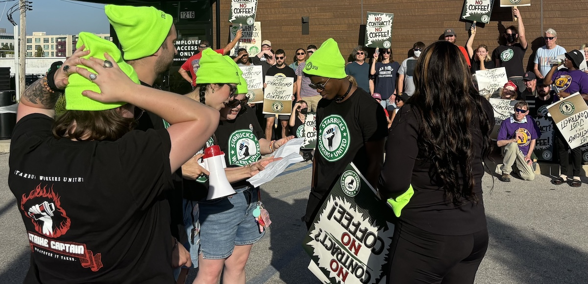 Several people in high-viz green stocking caps gather around a sheet of paper, pickets in the background have signs saying ‘no contract, no coffee’