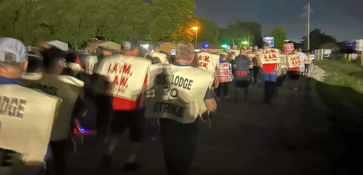 A crowd of workers walk away from the camera at night wearing tabards reading “District Lodge 70 on strike”