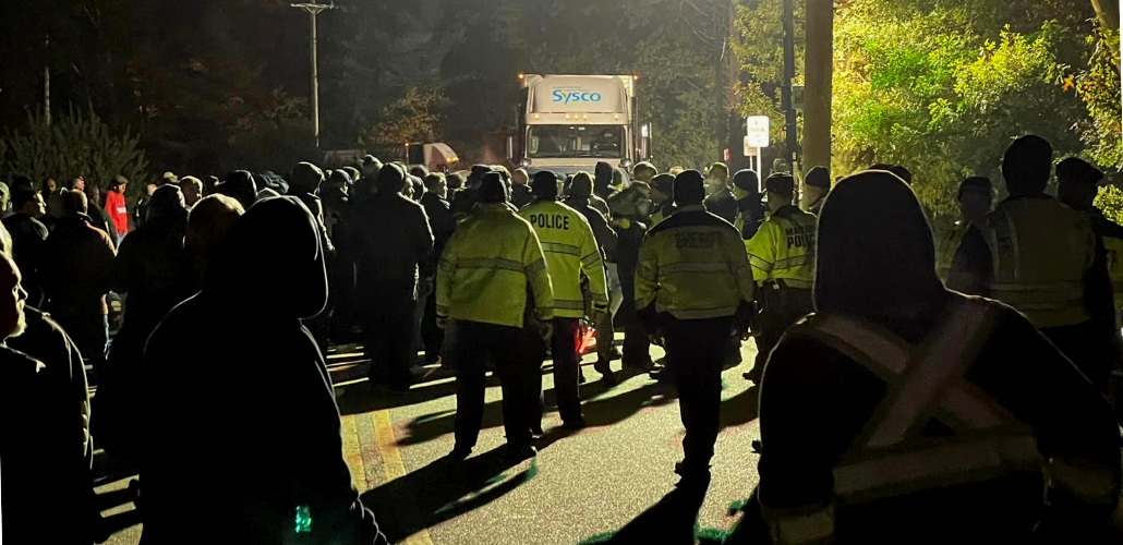 A Sysco truck faces off against a crowd of people, backs to the camera. A few wear yellow jackets that say "Police" or "Sheriff" on the backs; most do not. Around them it is still very dark out, but the central paved area is lit from above.
