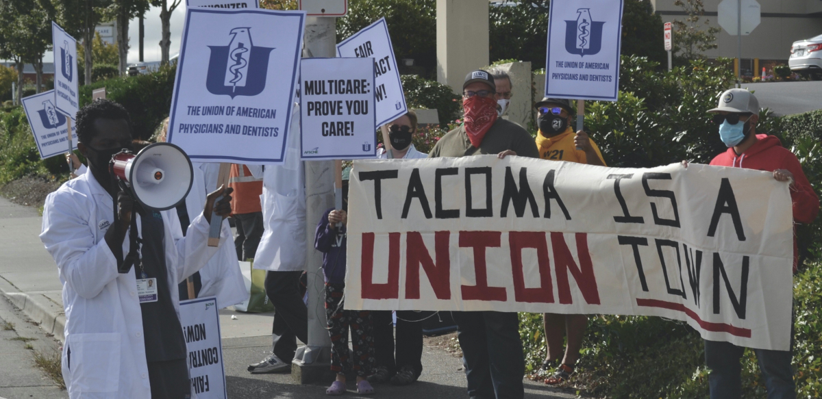 Urgent care doctors and staff in white coats picket alongside a sign held by union supporters that reads "Tacoma Is a Union Town."
