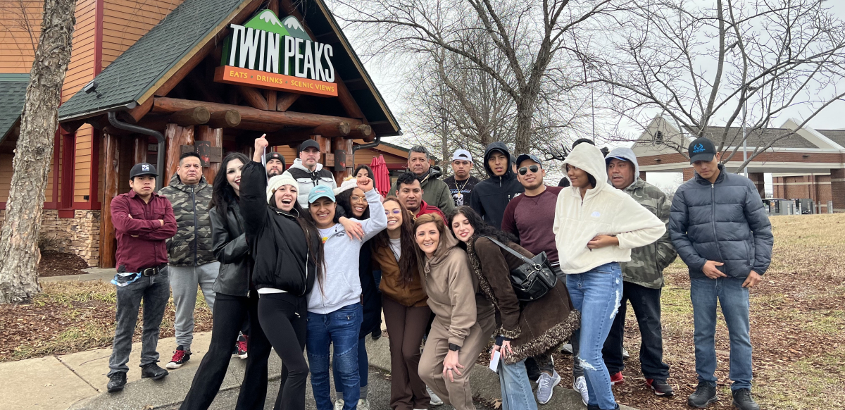 A racially diverse group of 21 workers, including women and men, poses outside in front of the Twin Peaks restaurant. Some have a tough and serious pose while others are smiling widely. Two have arms draped over each other's shoulders and fists in the air. The restaurant facade has a woodsy cabin style, with a logo of two mountains that may be meant to suggest breasts, and the slogan: "Twin Peaks: Eats, Drinks, Scenic Views."