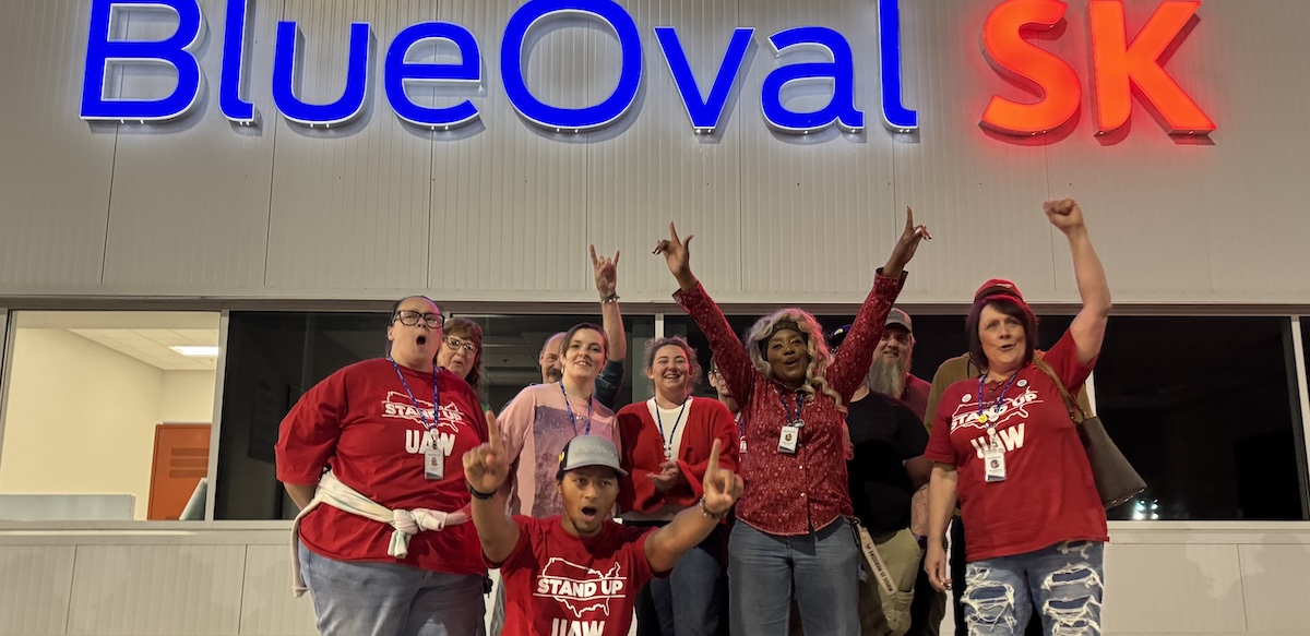 A group of 7 workers look excited and raise fists in front of a “BlueOval SK” sign