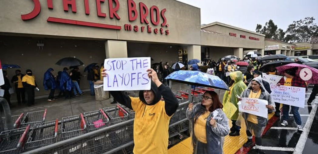 Workers in yellow shirts and rain parkas picket in front of a grocery store with a ‘stop the layoffs’ signs visible